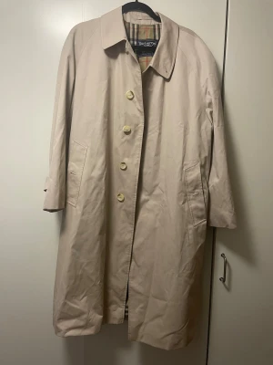Beige trenchcoat från Burberry - Endast använd ett par gånger, men tyvärr för stor. Rejält tyg. Klassisk beige trenchcoat från Burberry med det ikoniska rutiga fodret. Jackan har  knappar och raka fickor med knapp framtill. Perfekt för dig som vill ha en tidlös och stilren look. Som ni ser på en av bilderna: Etiketten i fickan står det ”52 short”.. Jag har fått den, men flera har bekräftat att den är äkta. Detta var innan de ändrade sitt namn till Burberry. Så det kan va vintage men är osäker. 