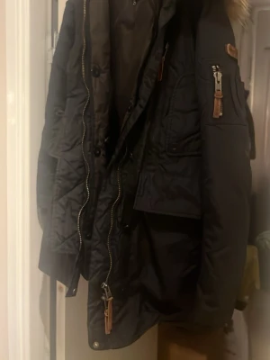 Svart parkas från MOSCOW - Säljer en svart parkas från MOSCOW  med päls på huvan, dragkedja och knappar framtill samt flera fickor med snygga detaljer i läderimitation. Jackan har en klassisk parkas-look och är perfekt för kalla dagar. Liten medium . 80% duckdown 20% feathers