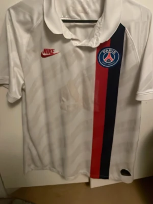 Vit PSG matchtröja från Nike - Säljer en vit PSG matchtröja från Nike med röd och marinblå rand på framsidan och klubbmärke på bröstet. Tröjan har korta ärmar, klassisk krage och diskreta Paris-tryck i tyget. Perfekt för dig som älskar fotboll och vill representera Paris Saint-Germain.