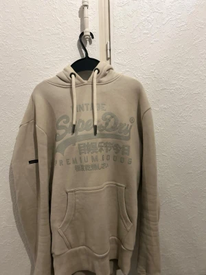 Beige hoodie från Superdry - Snygg beige hoodie från Superdry med stor logga och japansk text på bröstet. Hoodien har huva med dragsko, känguruficka och ribbade muddar. Perfekt för en avslappnad och trendig stil. Tillverkad i mjukt material för extra komfort.
