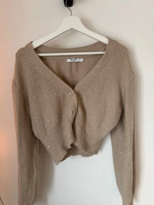 Beige glittrig kofta från NA-KD - Säljer en beige kofta från NA-KD med glittriga detaljer. Koftan har v-ringning, långa ärmar och knäpps med tre knappar framtill. Den är stickad och har en croppad passform som ger en trendig look.