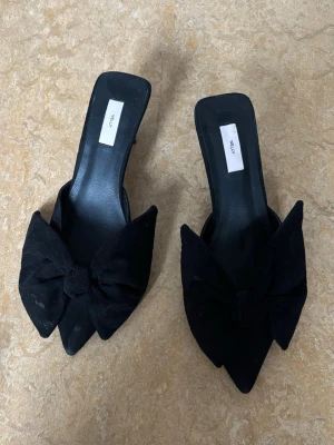 Svarta pumps med rosett från Nelly - Säljer ett par svarta pumps från Nelly med spetsig tå och stor rosett framtill. Skorna har låg klack och öppen häl, tillverkade i mockaimitation för en lyxig känsla. Perfekta för dig som vill ha en statement-detalj till outfiten. Klacken är 7cm