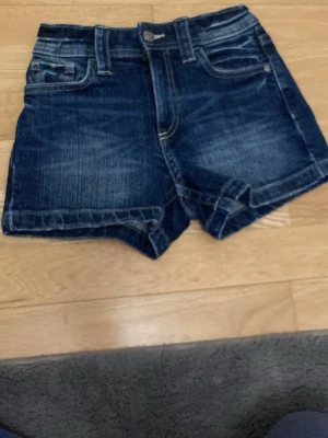 Mörkblå jeansshorts med fickor för barn  - Snygga mörkblå jeansshorts med klassisk femficksdesign och vikta benslut. Shortsen har bälteshällor, knapp- och dragkedjestängning samt dekorativa sömmar. Perfekta för varma dagar och enkla att matcha med olika toppar.