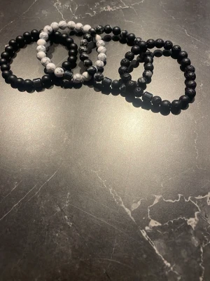 Set med svarta och vita pärlarmband - Fyra stilrena armband med pärlor i svart, vitt och marmorerat mönster. Elastiska band gör dem enkla att ta på och av. Perfekta för att addera en cool touch till din outfit. Materialet är troligen sten eller plast. Säljs som set.