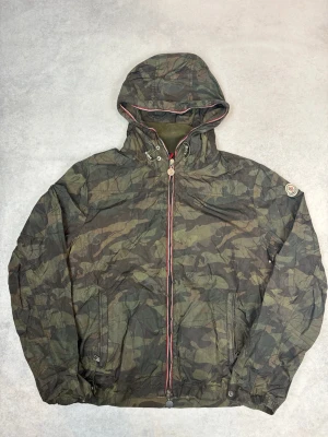 Camouflage vindjacka från Moncler - Snygg vindjacka från Moncler i camouflage-mönster med huva och hel dragkedja. Jackan har röda och vita detaljer längs dragkedjan och muddar vid ärmslut. Tillverkad i lätt och slitstarkt material, perfekt för blåsiga dagar. Moncler-logga på ärmen.