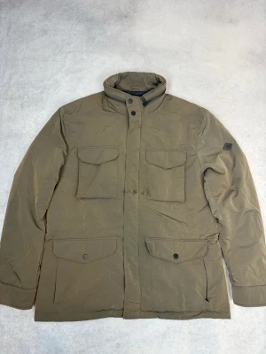 Olivgrön parkas från J.Lindeberg - Snygg olivgrön parkas från J.Lindeberg med fyra stora fickor framtill och hög krage. Jackan har dold dragkedja och tryckknappar, samt ett diskret märke på ärmen. Perfekt för kalla dagar med sin vadderade insida och klassiska design.