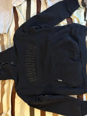 Svart hoodie från Hoodrich - Säljer en svart hoodie från Hoodrich med stor logga framtill och texten 'FROM NOTHING TO SOMETHING'. Hoodien har huva med dragsko och en stor magficka. Perfekt för dig som gillar streetwear och vill ha en clean look.