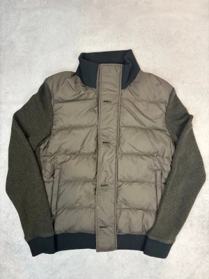 Brun pufferjacka med stickade ärmar från Woolrich - Snygg brun pufferjacka med quiltad front och rygg i glansigt material samt mörkare stickade ärmar. Jackan har hög ribbad krage, dragkedja och knappar framtill samt ribbade muddar. Perfekt för kalla vinterdagar.