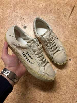 Saint Laurent vita sneakers - Snygga vita sneakers från Saint Laurent i canvas med handskriven logga på sidan. Skorna har lågt skaft, slitna detaljer och klassisk snörning. Perfekta för en avslappnad och trendig stil.