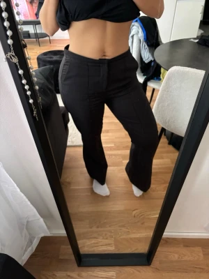 Svarta utsvängda kostymbyxor - Säljer ett par svarta kostymbyxor med utsvängda ben och hög midja. Byxorna har pressveck framtill och en klassisk, stilren look. Perfekta för dig som gillar en clean och modern stil.