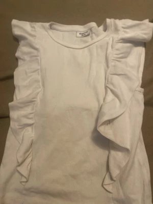 Vit volangtopp från Kappahl - Supersöt vit topp från Kappahl med volangdetaljer längs axlarna och sidorna. Tillverkad i ribbad bomull för en bekväm och mjuk känsla. Perfekt för dig som vill ha en enkel men ändå trendig look.