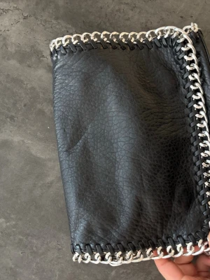 Svart kuvertväska med kedjedetaljer (från tiamo köpt på scorett) - Snygg svart kuvertväska i fejkläder med silverfärgade kedjedetaljer runt kanterna. Väskan har magnetlås och ett innerfack med dragkedja. Perfekt accessoar för att lyfta din outfit med en edgy touch.