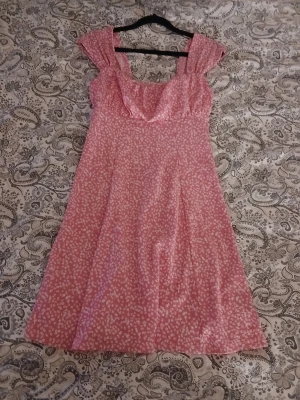 Rosa blommig klänning med volang - Söt rosa klänning med vita små blommor och volangdetalj vid bysten. Klänningen har breda axelband och en lätt utställd kjol som ger en feminin siluett. Perfekt för varma dagar och somriga tillfällen. Kan skicka fler bilder vid förfrågan 