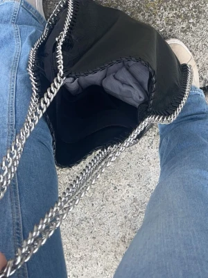 Svart kedjeväska  - Snygg svart handväska med silverfärgade kedjehandtag och dekorativa kedjor längs kanterna. Väskan har en mjuk, slouchy form och rymmer det viktigaste. Perfekt accessoar för dig som gillar en edgy och trendig look.