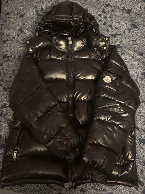 Svart dunjacka från Moncler med huva - Snygg svart dunjacka från Moncler med glansig finish, huva och dragkedja framtill. Jackan har Moncler-logga på ärmen, praktiska fickor med dragkedja och quiltad design. Perfekt för kalla dagar när du vill hålla stilen.