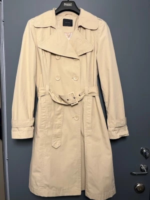 Vero Moda Trenchcoat – Elegant och tidlös i beige (Storlek L) - Upptäck den perfekta kappan för hösten och vintern! Denna klassiska trenchcoat från Vero Moda är både stilren och praktisk, med en elegant design som passar lika bra till jobbet som till en dag på stan.  Detaljer: Storlek: L Färg: Beige Stil: Klassisk trenchcoat med ett elegant snitt  Perfekt att bära över kavaj eller stickad tröja under kallare dagar Skick: Mycket fint - se bilder