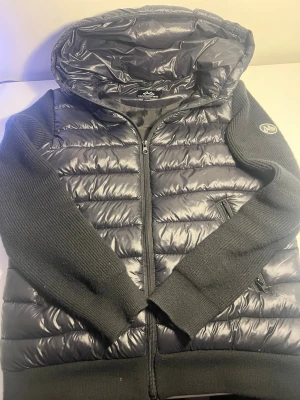 Svart pufferjacka från Maggiore - Snygg svart pufferjacka från Maggiore med stickade ärmar och huva. Jackan har quiltad front och rygg i glansigt material, dragkedja framtill och två sidofickor med dragkedja. Perfekt för kalla dagar och riktigt skön att bära. Riktigt fet! Stolek M i barn men är mer som Xs eller Xxs. Skicka gärna bud! Skick 9/10