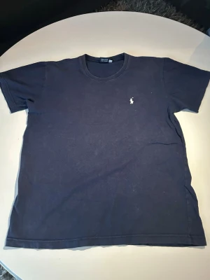 Mörkblå t-shirt från Ralph Lauren - Klassisk mörkblå t-shirt från Ralph Lauren med den ikoniska vita logon broderad på bröstet. T-shirten har rund halsringning och korta ärmar. Tillverkad i mjuk bomull för en skön och avslappnad stil.