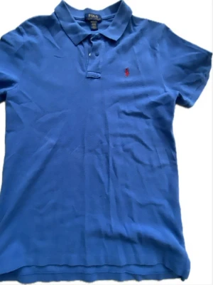 Blå pikétröja från Polo Ralph Lauren - Klassisk blå pikétröja från Polo Ralph Lauren med korta ärmar och krage. Tröjan har två knappar vid halsen och den ikoniska röda logotypen broderad på bröstet. Perfekt till jeans eller shorts för en avslappnad stil. 