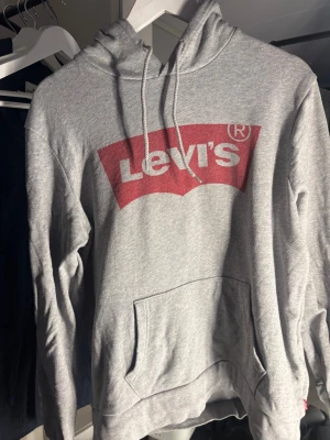 Grå Levi's hoodie med logga - Snygg grå hoodie från Levi's med stor röd och vit logga på bröstet. Klassisk känguruficka framtill och justerbar huva med dragsko. Tillverkad i mjukt bomullsmaterial som är perfekt för chill dagar eller streetwear-stil.