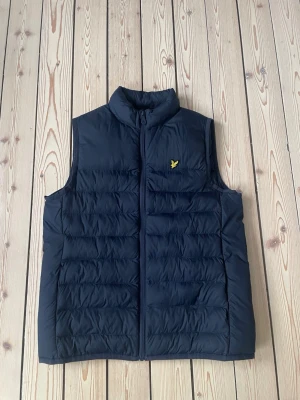 Mörkblå dunväst från Lyle & Scott - Snygg mörkblå dunväst från Lyle & Scott med gul logga på bröstet. Västen har hög krage, dragkedja framtill och quiltad design. Perfekt för lager-på-lager och enkel att matcha med olika outfits.