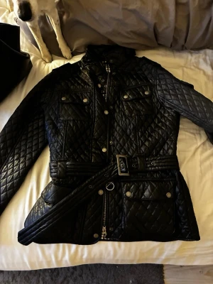 Svart quiltad jacka med bälte - Snygg svart quiltad jacka med bälte i midjan och flera fickor framtill. Jackan har hög krage, dragkedja och tryckknappar samt dekorativa sömmar i rutmönster. Perfekt för dig som vill ha en stilren och trendig look.