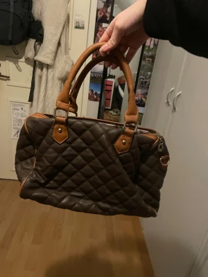 Brun  handväska  - Snygg brun handväska i quiltat mönster med ljusbruna detaljer och dubbla handtag. Väskan har dragkedja upptill och ett blommigt innerfoder. Perfekt storlek för att få plats med allt du behöver.