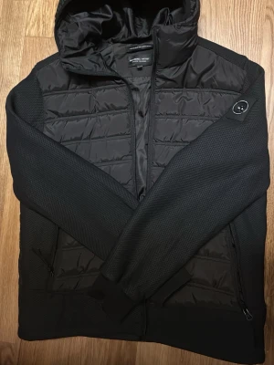 Svart jacka från Marshall Artist - Snygg svart jacka från Marshall Artist med quiltad front och stickade ärmar. Jackan har huva, dragkedja och två fickor med dragkedja. Perfekt för dig som gillar streetwear och stilren design.