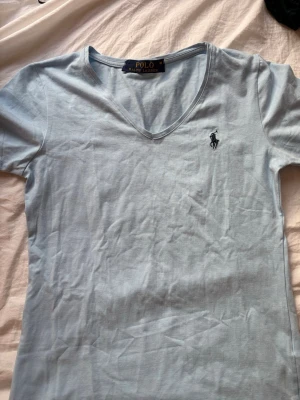 Ljusblå t-shirt från Polo Ralph Lauren - Snygg ljusblå t-shirt från Polo Ralph Lauren med v-ringning och den klassiska broderade loggan på bröstet. Tillverkad i mjukt bomullsmaterial och har en enkel, stilren design som passar till det mesta.