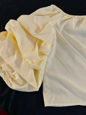 Gul offshoulder blus med puffärm - Trendig gul offshoulder blus med en stor puffärm på ena sidan. Tillverkad i ett lätt, vävt material som ger en luftig känsla. Perfekt för dig som vill sticka ut med en unik och cool look.