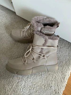 Beige moon boots med fuskpäls 40 oanvänd  - Säljer ett par beige moon boots med fluffig fuskpäls runt skaftet och grov sula. Skorna har snörning framtill och dragkedja på sidan för enkel påtagning. Perfekta för kalla dagar och ger en trendig vinterlook.