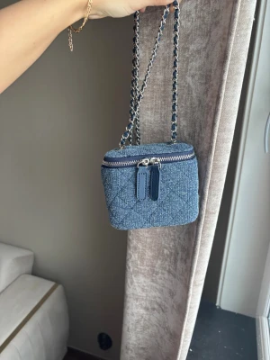 Blå quiltad axelväska i denim - Trendig blå axelväska i quiltad denim med silverfärgad dragkedja och kedjerem med insytt tyg. Väskan har boxig form och dubbla dragkedjor framtill. Perfekt för dig som gillar en cool och modern look.