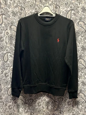 Svart sweatshirt från Polo Ralph Lauren - Klassisk svart sweatshirt från Polo Ralph Lauren med rund halsringning och det ikoniska röda broderade logot på bröstet. Tröjan har ribbade muddar vid ärmslut och nederkant. Perfekt för en stilren och avslappnad look.