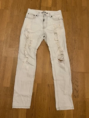 Adrian Hammaond jeans - strl.W31 - Säljer ett par ljusa jeans med flera slitningar och hål på både fram- och baksida. Modellen är rak och har fem fickor samt bälteshällor. Jeansen har en avslappnad vibe och passar perfekt till streetwear-stilen.