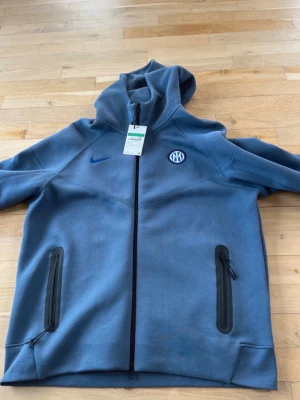 Blå Inter Nike tech - Snygg blå hoodie från Nike med Inter-logga på bröstet och blå Nike Swoosh. Tröjan har hel dragkedja framtill, två fickor med dragkedja och en stor huva. Perfekt för dig som gillar sportig stil och vill visa ditt stöd för Inter. Köpt för 141 euro mitt pris 600. Helt ny