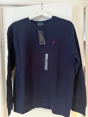 Mörkblå sweatshirt från Polo Ralph Lauren - Klassisk mörkblå sweatshirt från Polo Ralph Lauren med rund halsringning och det ikoniska röda broderade logot på bröstet. Tröjan har långa ärmar och ribbade muddar vid ärmslut och nederkant. Perfekt för en stilren och avslappnad look. Nypris 1195kr 