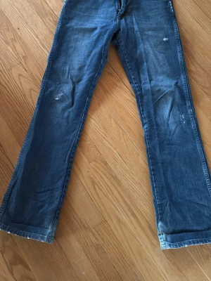Wrangler Blå bootcut jeans med slitningar - Säljer ett par klassiska blå bootcut jeans med lätt slitna detaljer på benen. Jeansen har normal midja och är tillverkade i denim med en snygg tvätt. Stl 30/34
