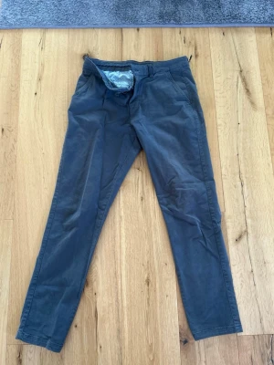 Grå/ blågrå chinos med smal passform - Snygga grå/ gråblå chinos med slimfit passform och klassisk design. Byxorna har två sidofickor fram och två bakfickor med knapp. Tillverkade i mjukt bomullstyg som ger en skön känsla hela dagen.