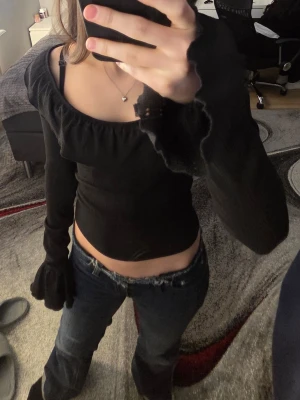 Svart croppad topp med volang - Trendig svart croppad topp med långa ärmar och volangdetalj längs urringningen. Toppen har en figurnära passform och är perfekt för att lyfta vilken outfit som helst. Materialet känns mjukt och stretchigt, vilket gör den bekväm att bära.