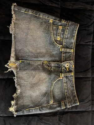 Blå jeans kjol med rå kant och ljus gröna detaljer ! - Snygg blå jeans kjol med slitna, råa kanter och ljus grön kontrastsöm. Klassisk femficksmodell med dragkedja och knapp. Bakfickorna har dekorativa sömmar. 