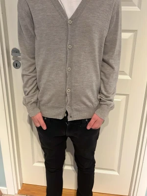 Ljusgrå kofta från j Lindeberg - Säljer en stilren ljusgrå kofta från j. Lindeberg med v-ringning och knappar framtill. Tillverkad i mjuk bomull och har ribbade muddar vid ärmslut och nederkant. Perfekt att slänga över en t-shirt för en clean look.