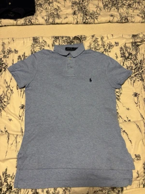 Ljusblå pikétröja från Ralph Lauren - Snygg ljusblå pikétröja från Ralph Lauren med klassisk krage och knappslå. Tröjan har korta ärmar och den ikoniska broderade loggan på bröstet. Perfekt för en stilren och avslappnad look.