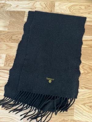 Svart halsduk från Barbour i ull - Klassisk svart halsduk från Barbour i mjuk ull med broderad logga i gult. Halsduken har fransar i båda ändar och en enkel, stilren design som passar till det mesta. Perfekt accessoar för kyliga dagar.