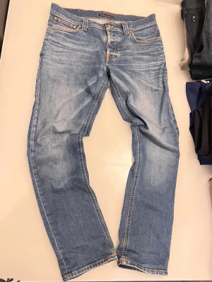 Blå jeans från Nudie Jeans - Säljer ett par klassiska blå jeans från Nudie Jeans med snygg tvätt och kontrastsömmar. Modellen har fem fickor, normal midja och raka ben. Materialet är mjukt denim i bomull och bakfickan har Nudie Jeans signaturbrodyr.