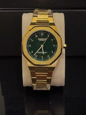 Klocka Kompass  - Stilren guldfärgad klocka från Kompass med grön urtavla och arabiska siffror. Klockan har en länk i metall och en klassisk, kantig design som ger en lyxig känsla. Perfekt accessoar för att lyfta din outfit.