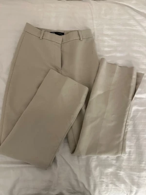  Kostymbyxor med raka ben - Snygga beigea kostymbyxor med klassisk passform och raka ben. Byxorna har hällor för bälte, diskreta sidofickor och en bakficka med passpoal. Perfekta för en stilren och avslappnad look. Materialet känns mjukt och lätt, vilket gör dem bekväma att bära hela dagen.