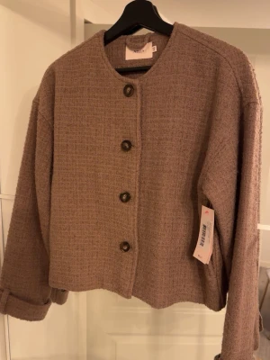 Beige cardiganjacka från Nelly - Stilren beige cardiganjacka från Nelly med rund halsringning och fyra bruna knappar framtill. Jackan har en rak, boxig passform och är gjord i ett mjukt, strukturerat tyg som ger en trendig look. Perfekt att slänga över en t-shirt eller topp för extra stil. Passar både XS och S