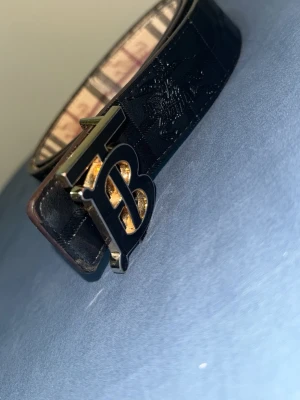 Svart Burberry bälte med B-spänne - Snyggt svart bälte från Burberry med ett stort B-format metallspänne i svart och guld. Bältet har präglad logga och klassiskt rutigt mönster på insidan. Perfekt accessoar för att lyfta din outfit.