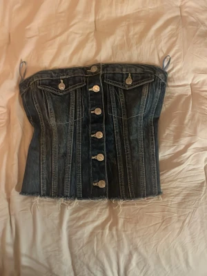 Mörkblå jeanskorsett med knappar - Säljer en snygg mörkblå korsettopp i jeans med dekorativa sömmar och silverfärgade knappar framtill. Toppen har smala axelband och rå kant nertill för en edgy look. Perfekt att styla med höga byxor eller kjol.