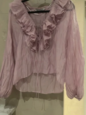 Ljuslila volangblus från Zara - Supersöt ljuslila blus från Zara med stora volanger längs ringningen och knytband framtill. Blusen är gjord i ett tunt, transparent tyg och har långa, puffiga ärmar med resår vid handlederna. Perfekt för dig som gillar romantisk och trendig stil.